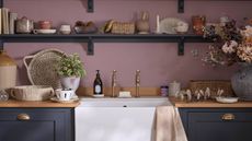 A purple laundry room with dark navy shelving and cabinets, and a large white sink with gold faucets. On the shelves are trays, trinkets, and a large grey vase.