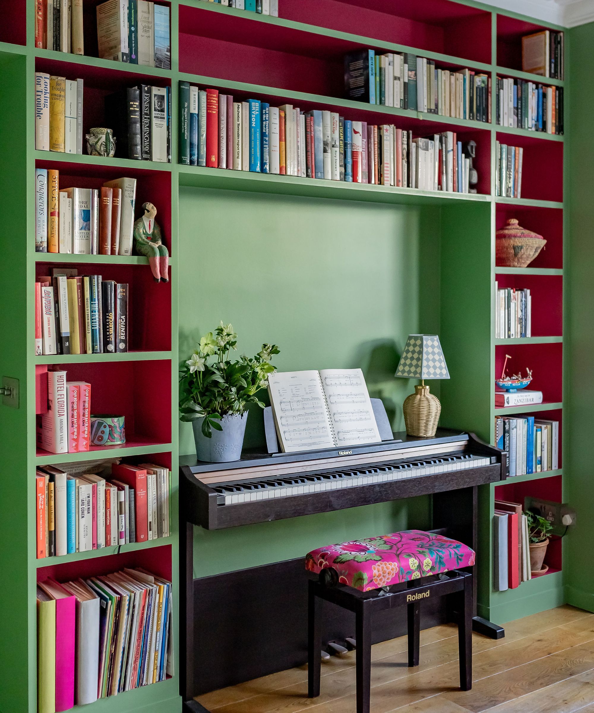 Living room with green and pink painted built-in bookcases filled with books, a piano with a plant and lamp on top, and a stool with a floral cushion on top.