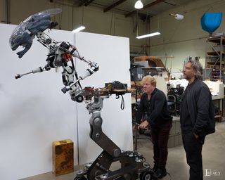 Two men stand beside a robotic alien monster at a studio