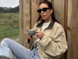 British fashion influencer Jessica Skye at a cafe in Cornwall holding a mug while wearing black oval sunglasses, silver earrings, ivory cable knit sweater, cropped barn coat, black studded Khaite belt, and jeans