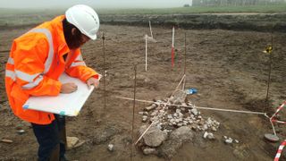 an archaeologist in an orange jacket bent over a tablet and sketching. beside them on the ground is a set of stones in an approximatel circular shape. the area is marked off and protected by small poles and strings 