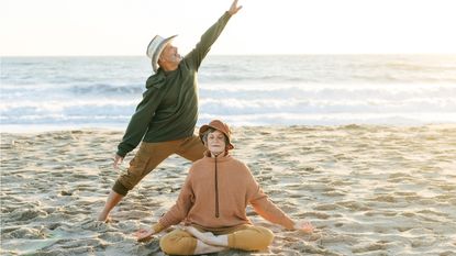 A retired couple do yoga poses on a beach.