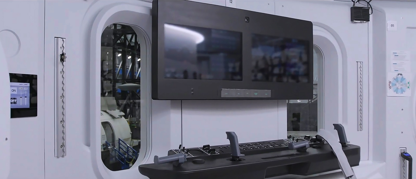 a joystick instrument panel sits under two black screens, with two tall oval windows on either side, inside a white room.
