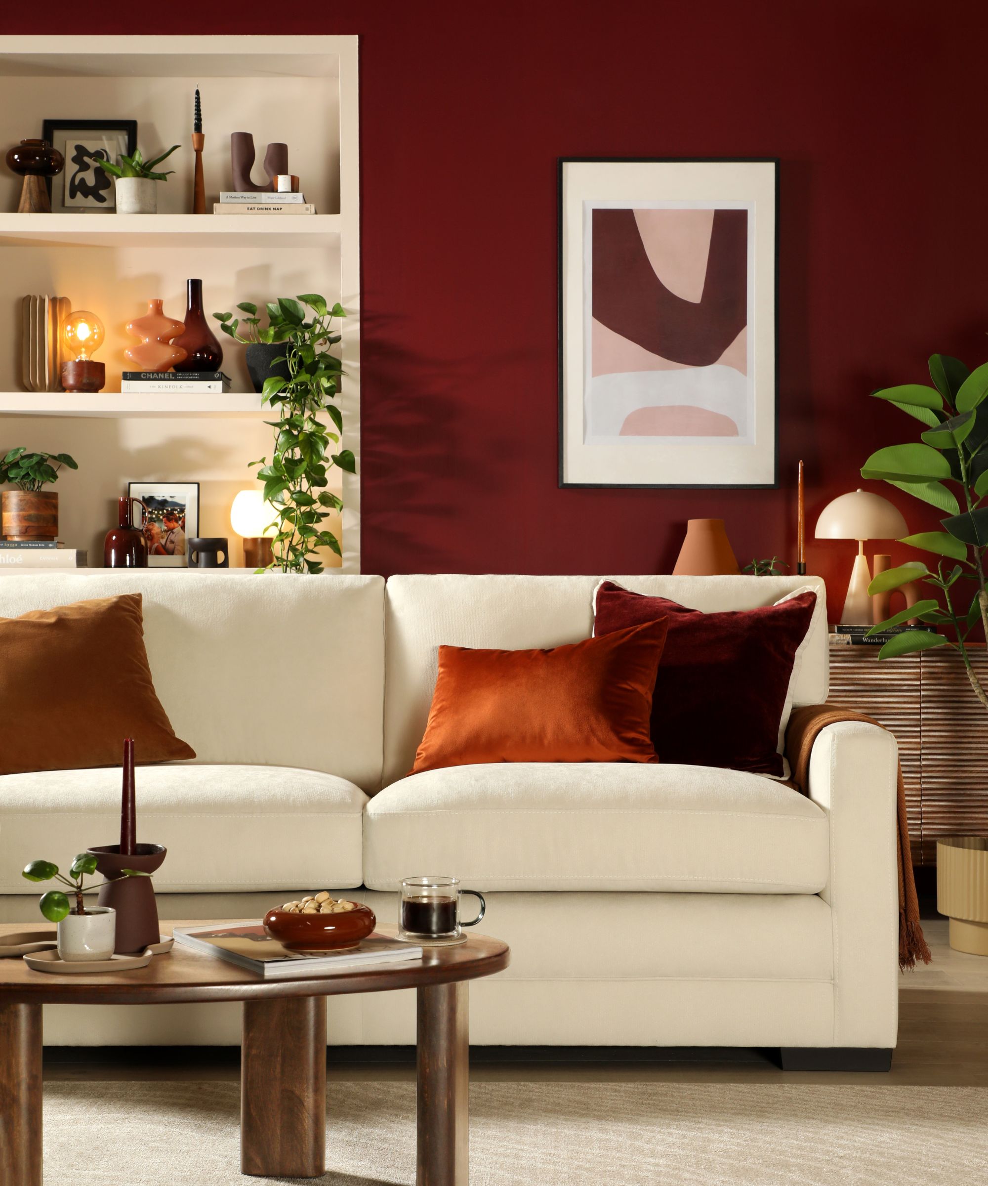 A deep crimson living room with a cream sofa layered with burnt orange and burgundy velvet cushions, a round walnut coffee table, styled bookshelves and abstract framed artwork above.
