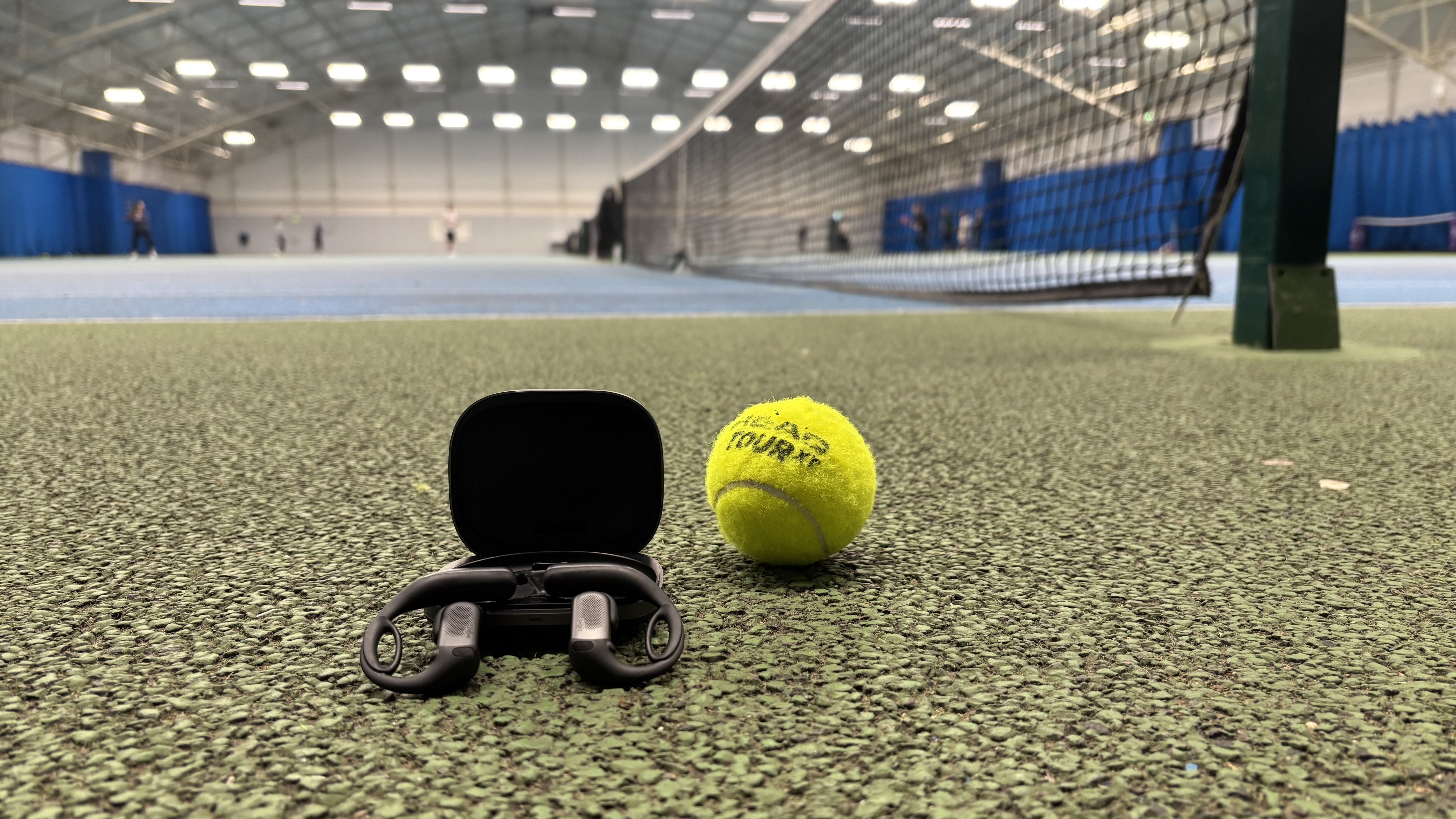 Shokz OpenFit Pro open earbuds, balancing on their carry case, next to a tennis ball at an indoor tennis center.