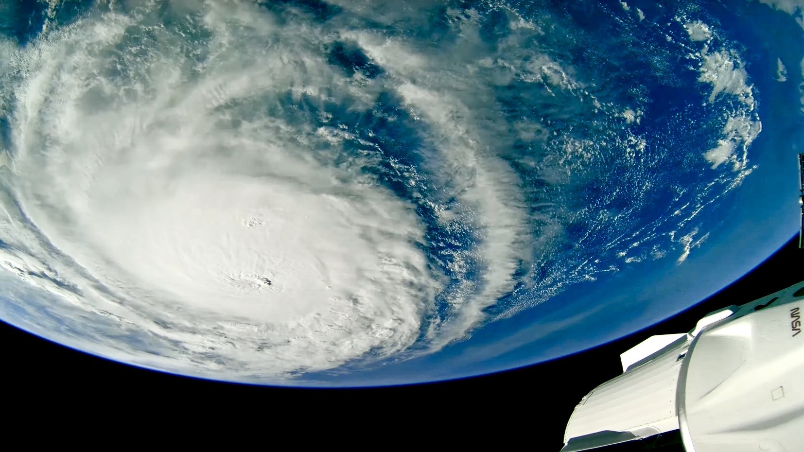 The bulbous blue orb of Earth hangs from above. On its face, on the left, a giant white swirl of this cloud, a hurricane. On the bottom right, within the sliver of black space beneath the Earth, a white spacecraft is partially seen.