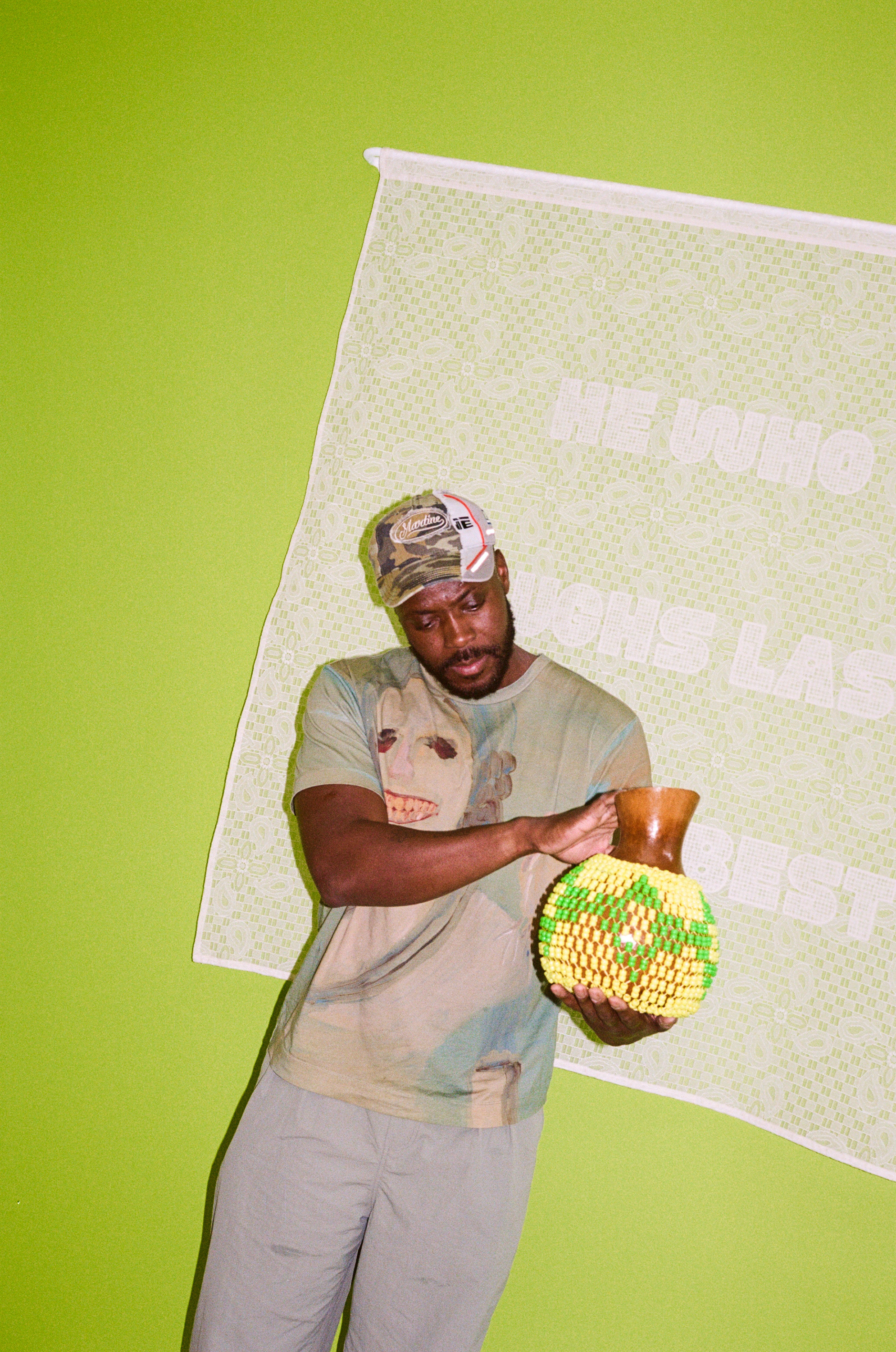 Yinka Ilori holding a custom-made shekere decorated with beads, against green wall