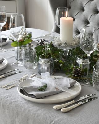 Christmas table setting with glass candlestick with pillar candle and foliage around it, glassware, tealights, white plates, grey linen tablecloth