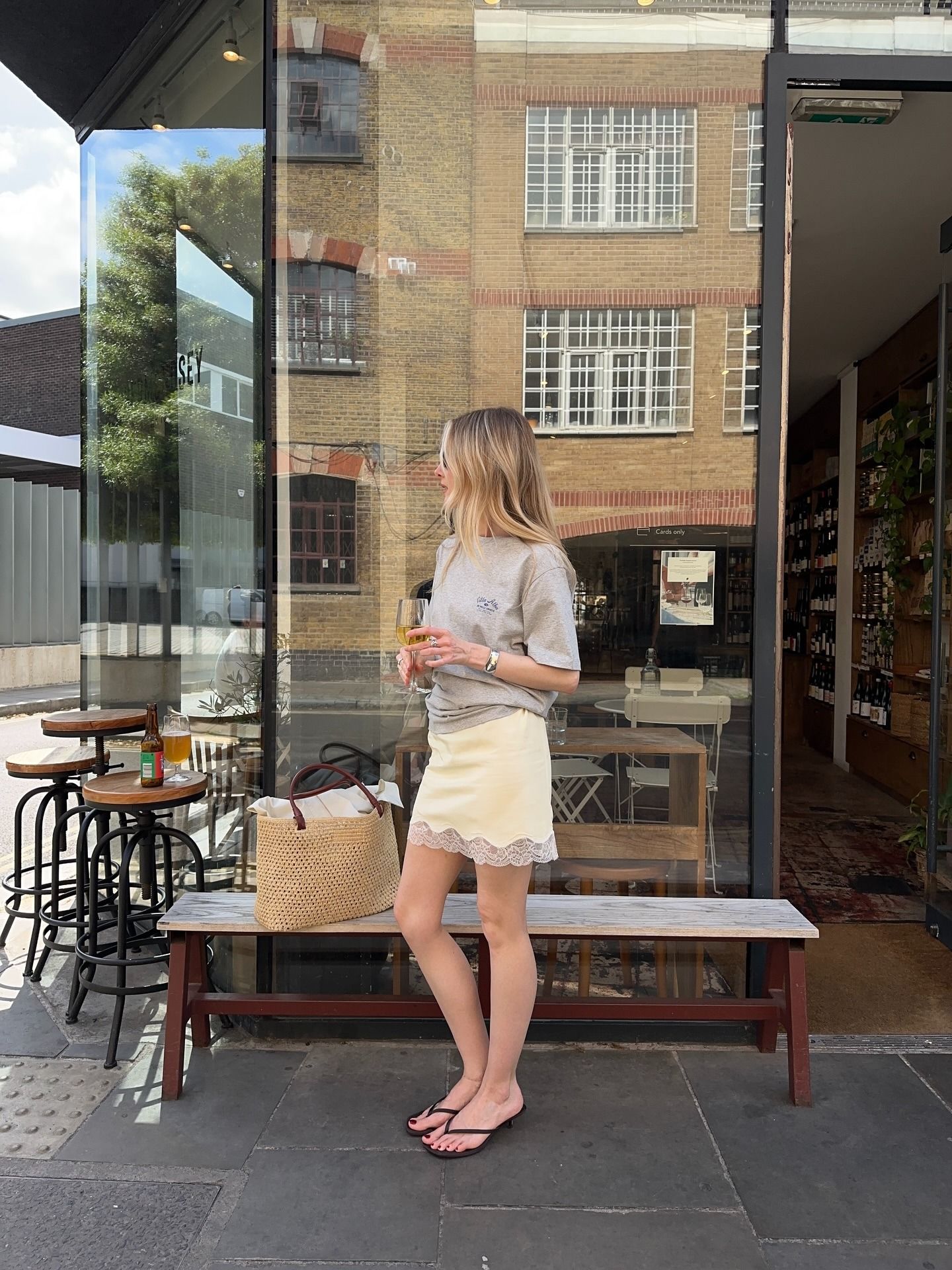 chloe wears a yellow skirt, raffia tote, flip flops and a gray tee.