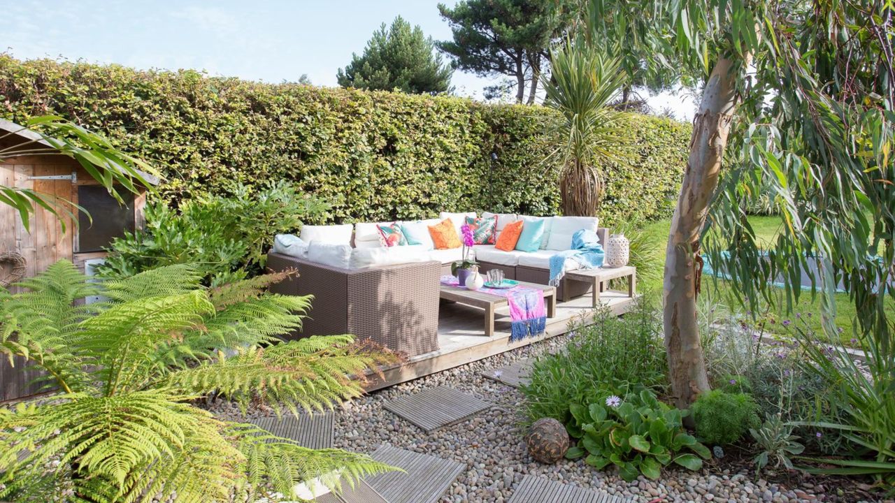 Sofa on a decked area beside a hedge, a gravelled area and lawn. The garden of a 1970s house, home of Alison and Douglas Gibb and their two children.