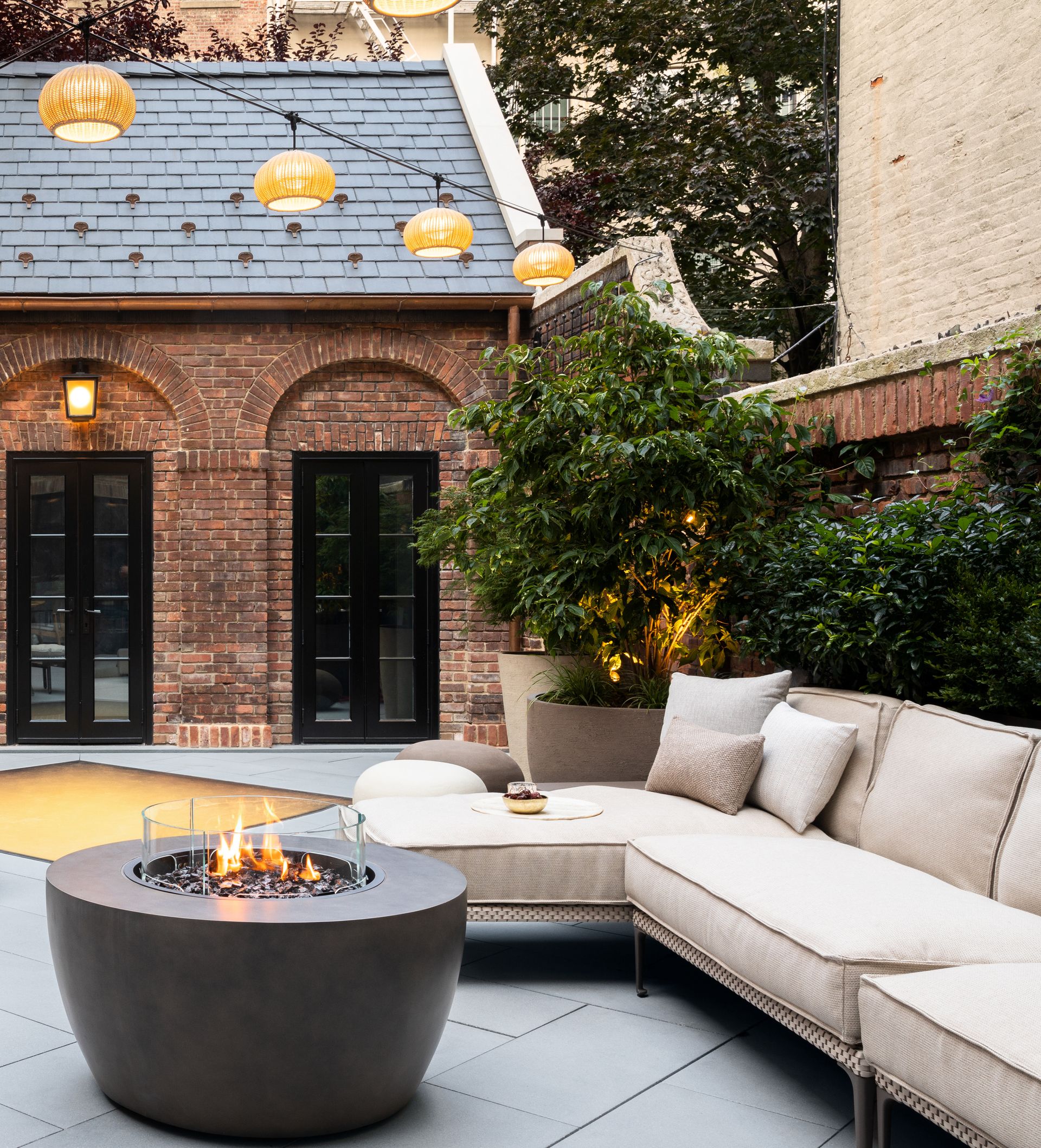 An outdoor space with plants along the back wall, a sofa complete with firepit lit during the day, and hanging lanterns
