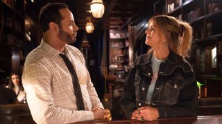 DANIEL SUNJATA and KAITLIN OLSON as Karadec and Morgan looking at each other while leaning against a table in High Potential.