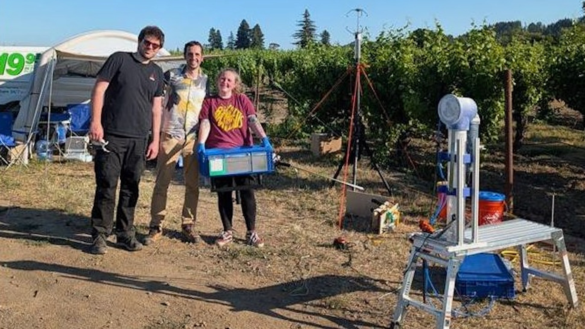 three people stand near a metallic piece of scientific gear outside, with trees in the background