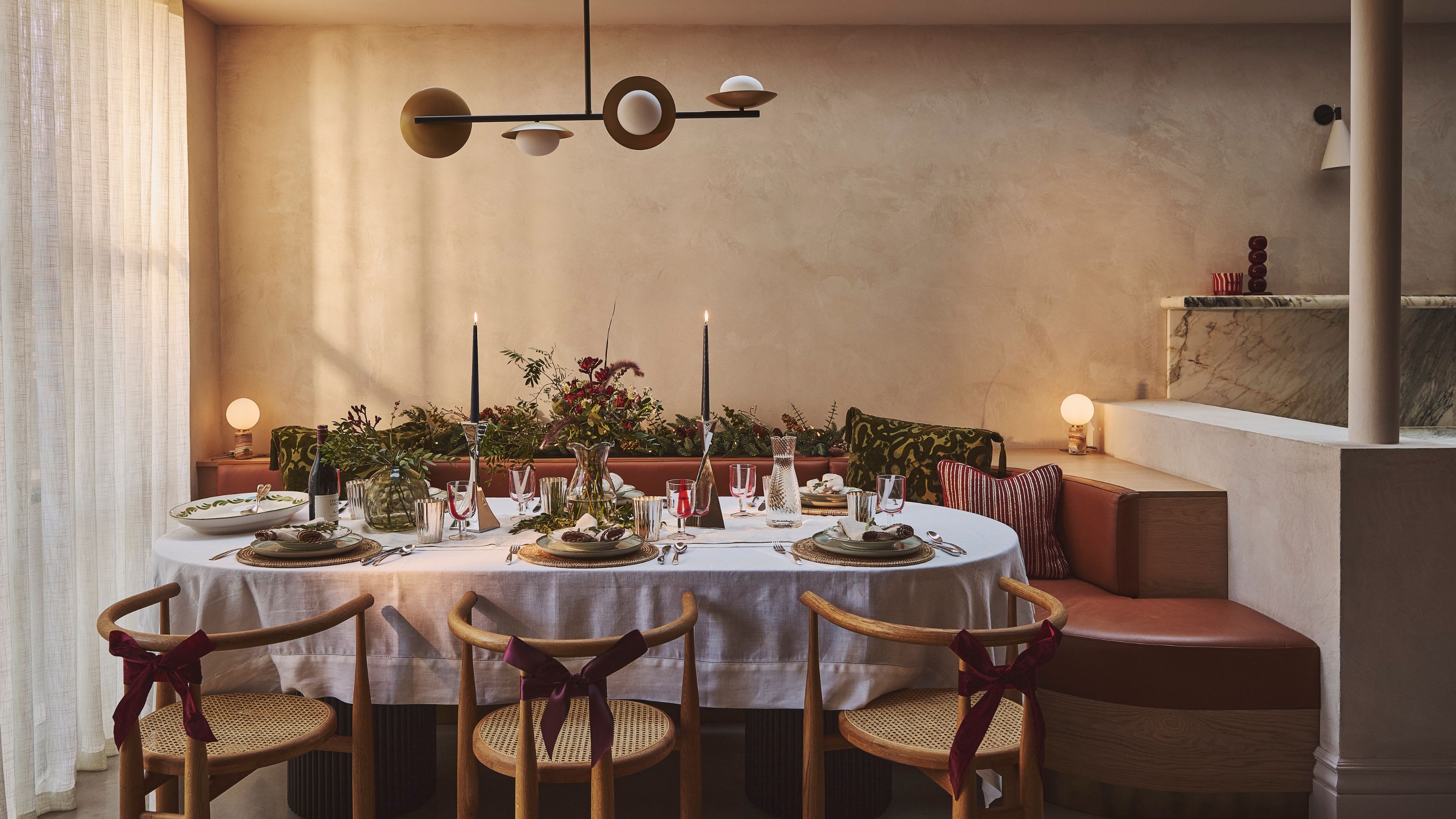 A modern dining room with a festive tablescape with a white tablecloth and long candles, wooden dining chairs and banquette seating. 
