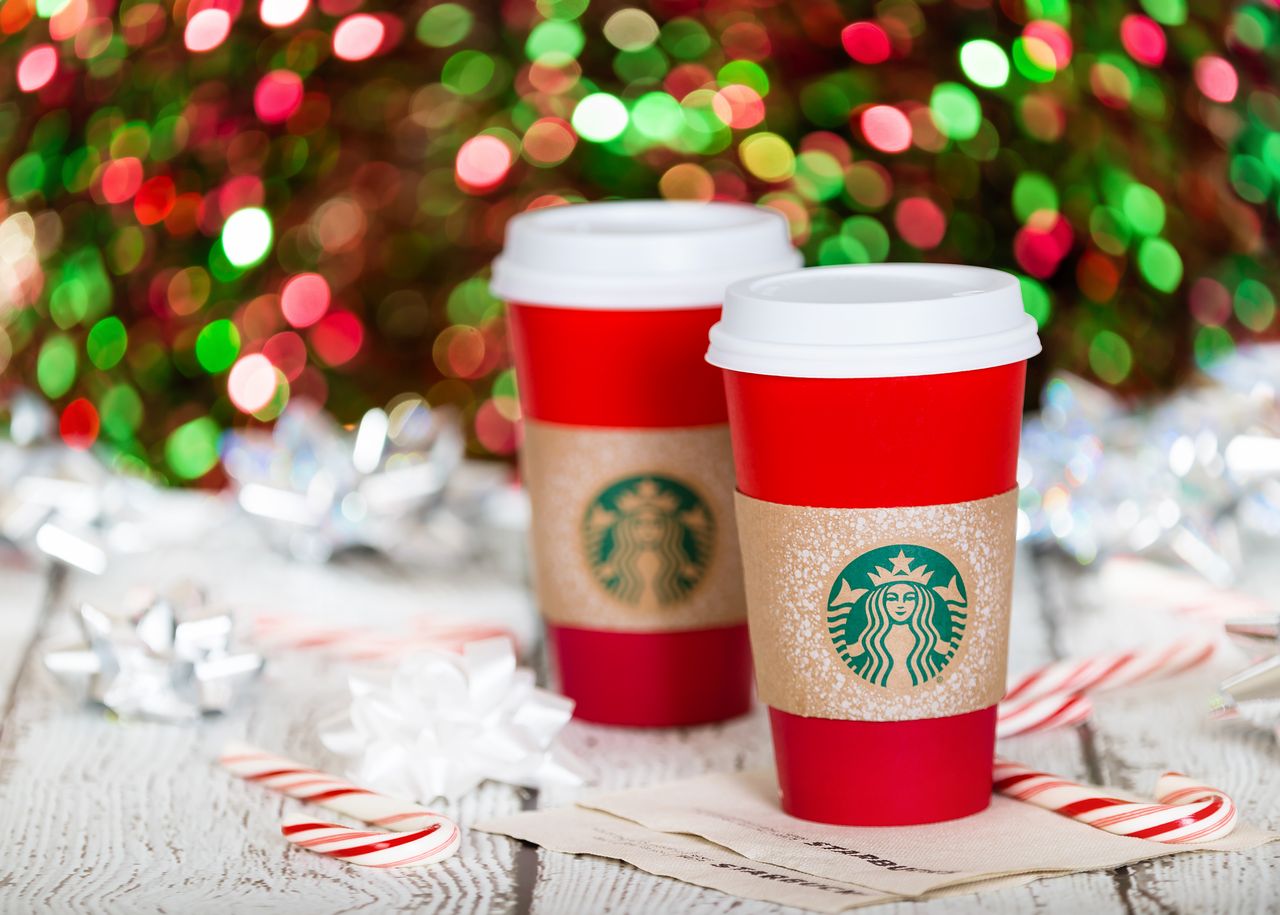 Christmas tree and lights in the background and two holiday Starbucks cups on a table with a candy cane.