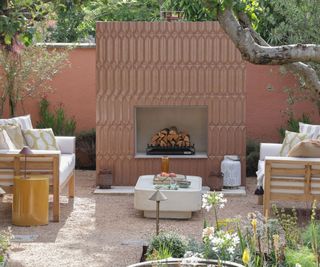 A terracotta-toned garden with an outdoor fireplace clad in textured 3D relief tiles, flanked by white cushioned timber sofas, a concrete coffee table and gravel ground. 