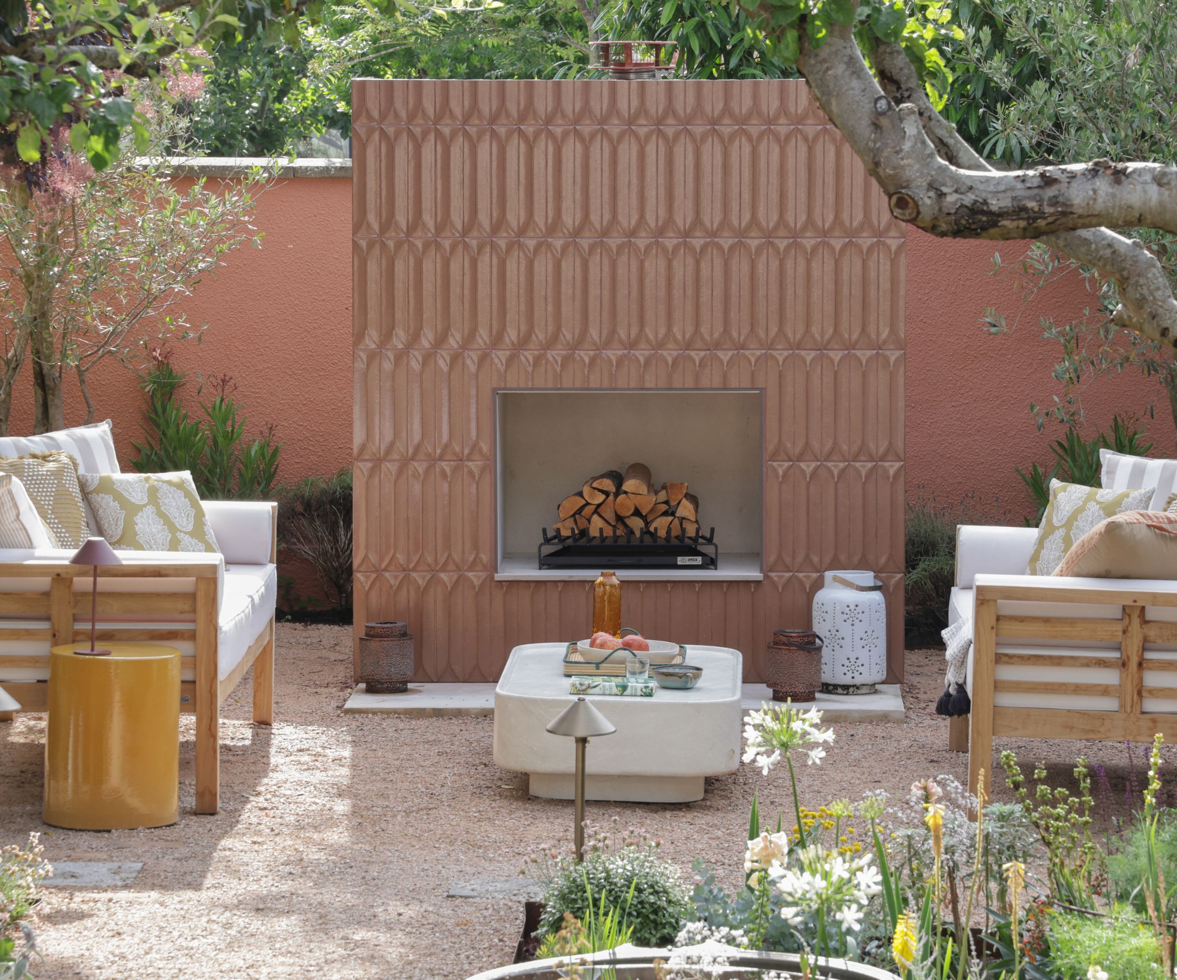 A terracotta-toned garden with an outdoor fireplace clad in textured 3D relief tiles, flanked by white cushioned timber sofas, a concrete coffee table and gravel ground.