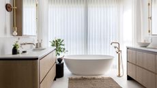 White bathroom with voile blinds behind tub and rattan rug and plant