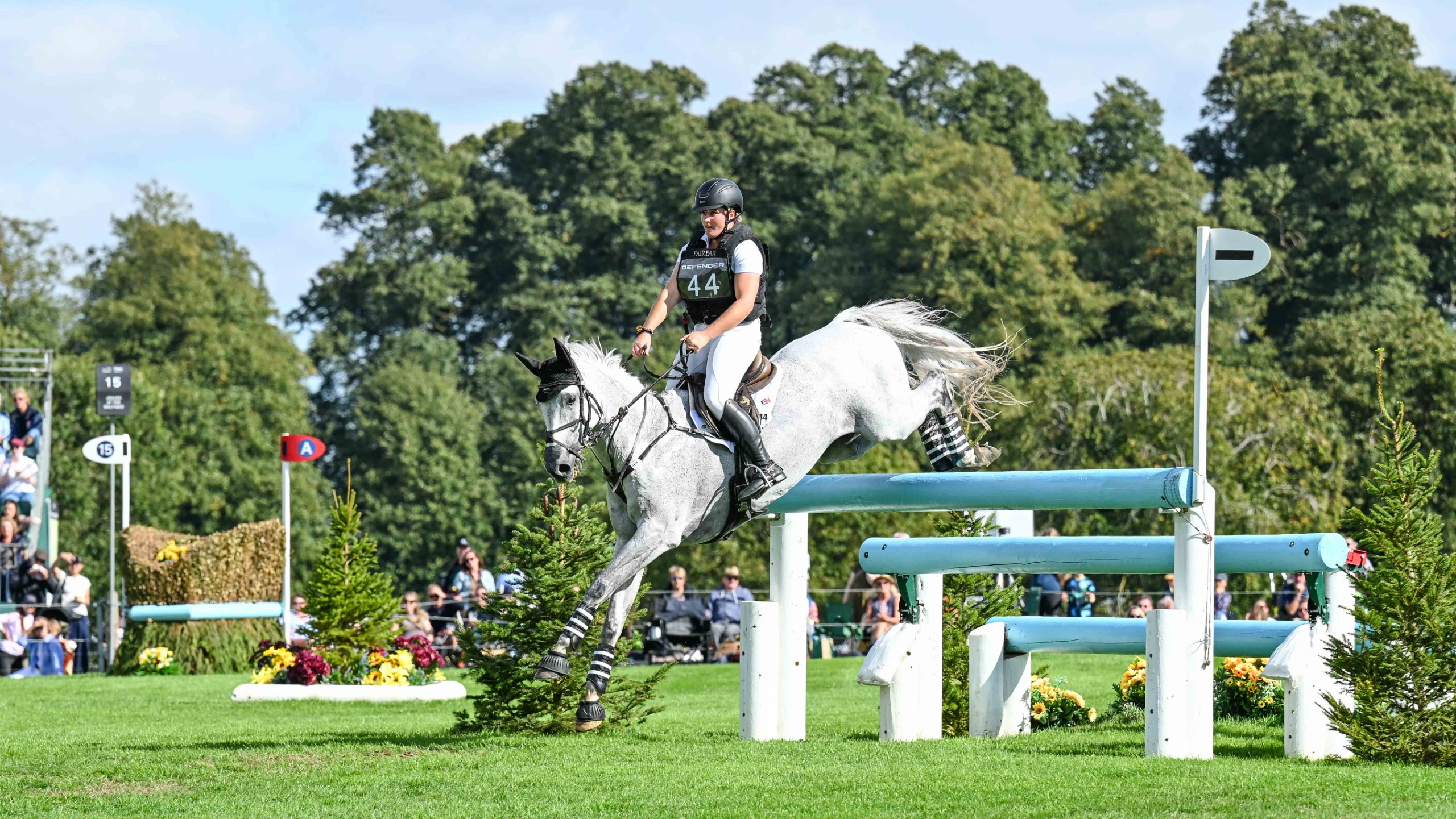 Ema Klugman wearing a white top and a black body protector and hat clears an obstacle on her grey horse Chiraz at the 2026 Defender Burghley Horse Trials 