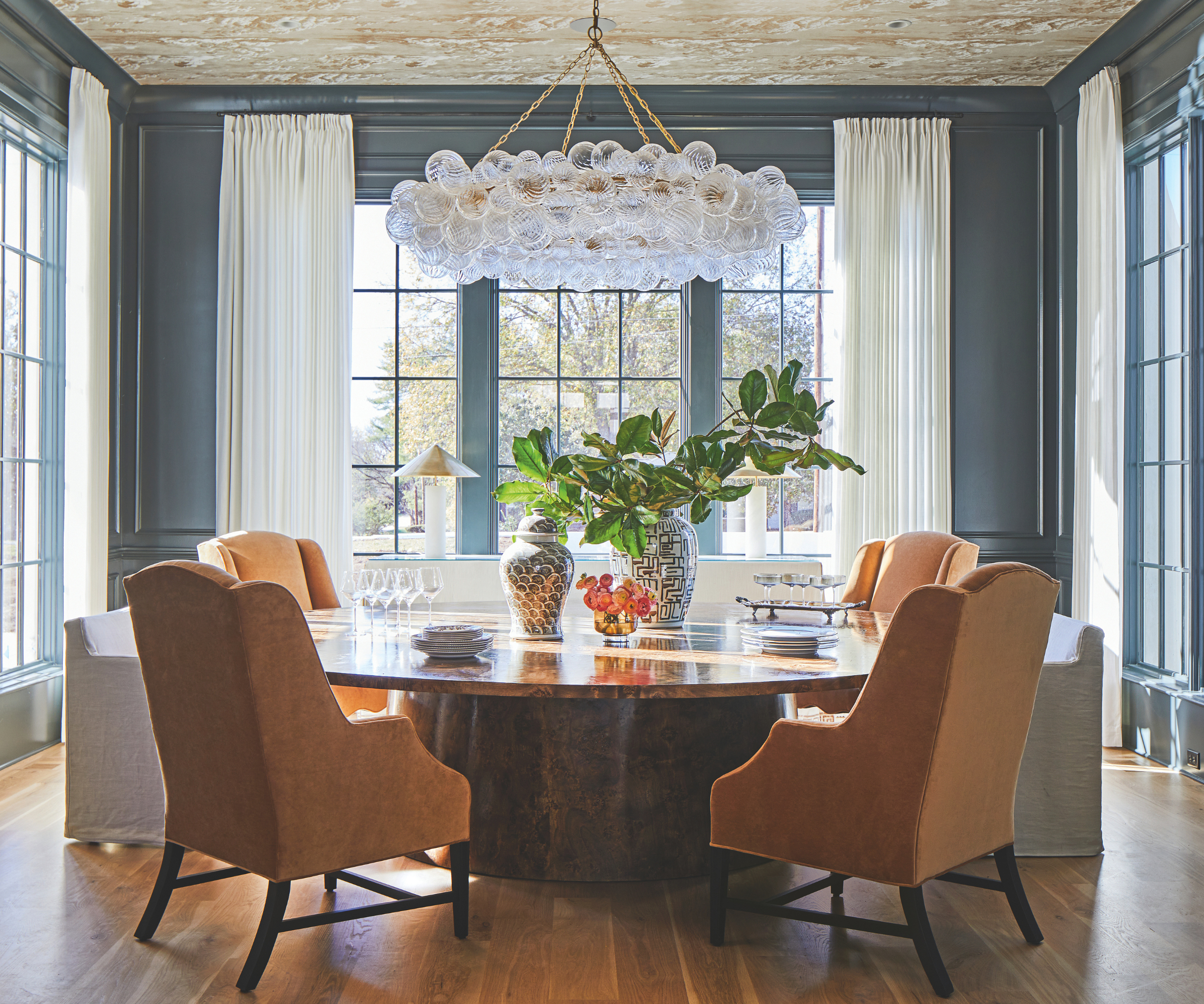 Blue dining room with large windows and white drapes. A large polished wood circular dining table sits in the middle with orange upholstered dining chairs around