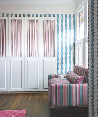 A bright and patterned bedroom with wooden flooring, pink rug, striped sofa, white and blue patterned wallpaper and a white closet with pink curtains. Large windows with white shutters flood the room with light.