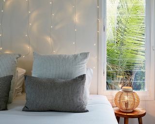 A bedroom with striped pillows, curtain light behind bed, a white rectangular window and a bedroom side light