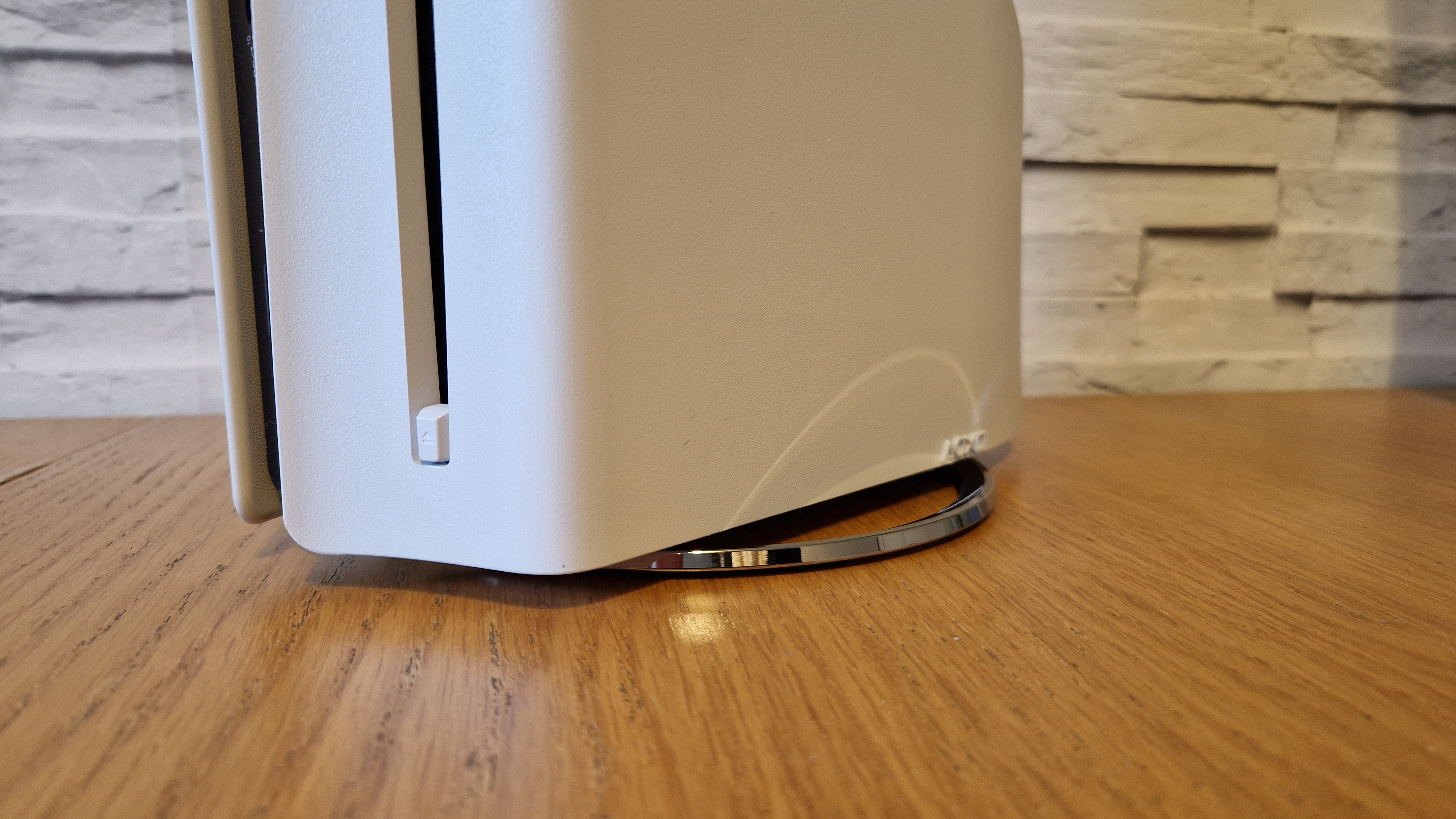 A close up of the PS5 Slim's vertical stand on a wooden table with a white brick background