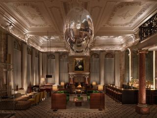 A grand hotel lobby features renaissance style architecture contrasted with modern furnishings in textile and wood and a colossal, mirrored oval sculpture hanging from the ceiling.