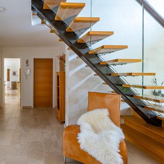 Tiled hallway with beautiful wooden staircase and a glass railing leading the way upwards next to a leather chair