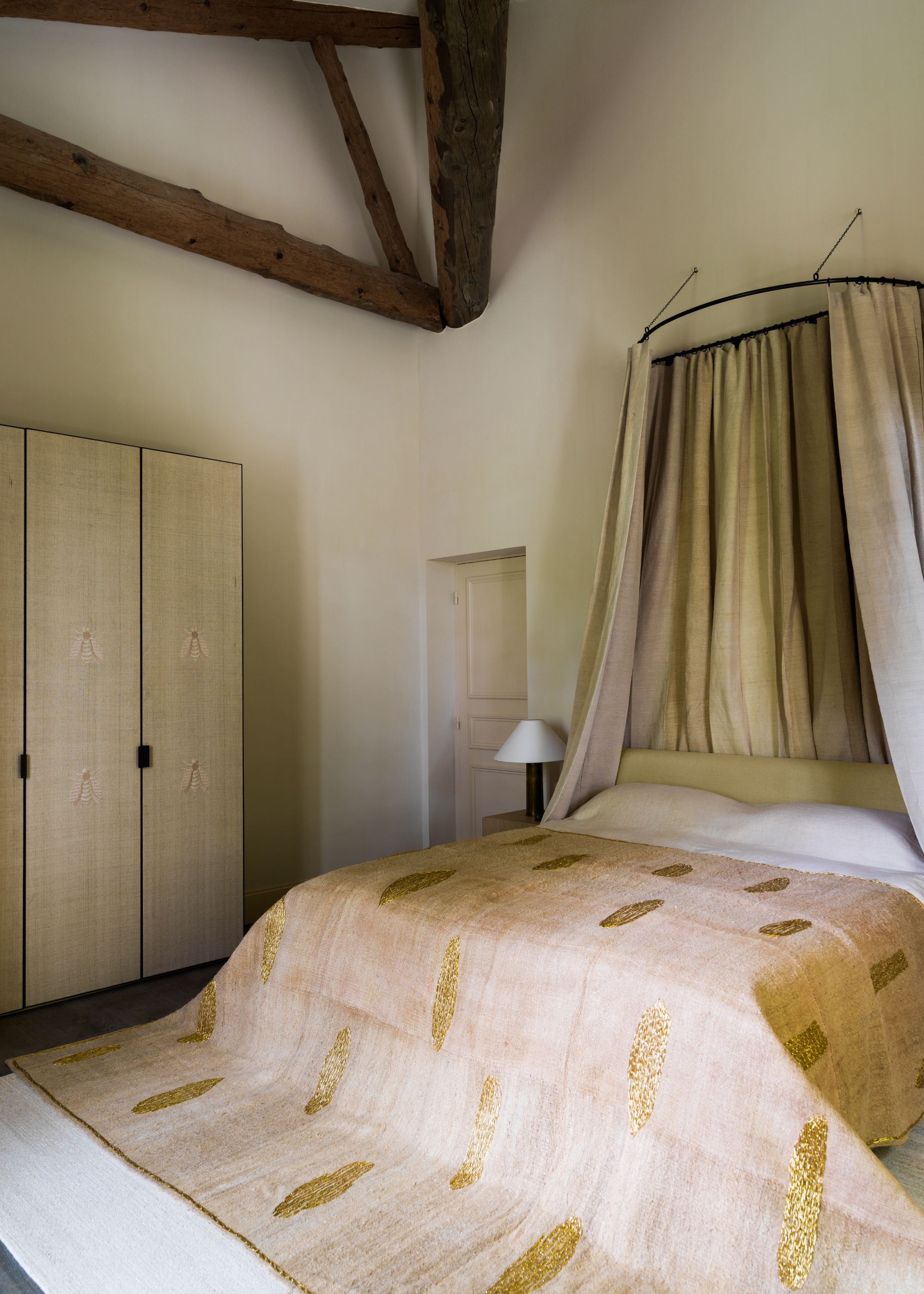 A bedroom with a bed canopy, a patterned duvet, white pillows, a table lamp, and a minimalist wardrobe