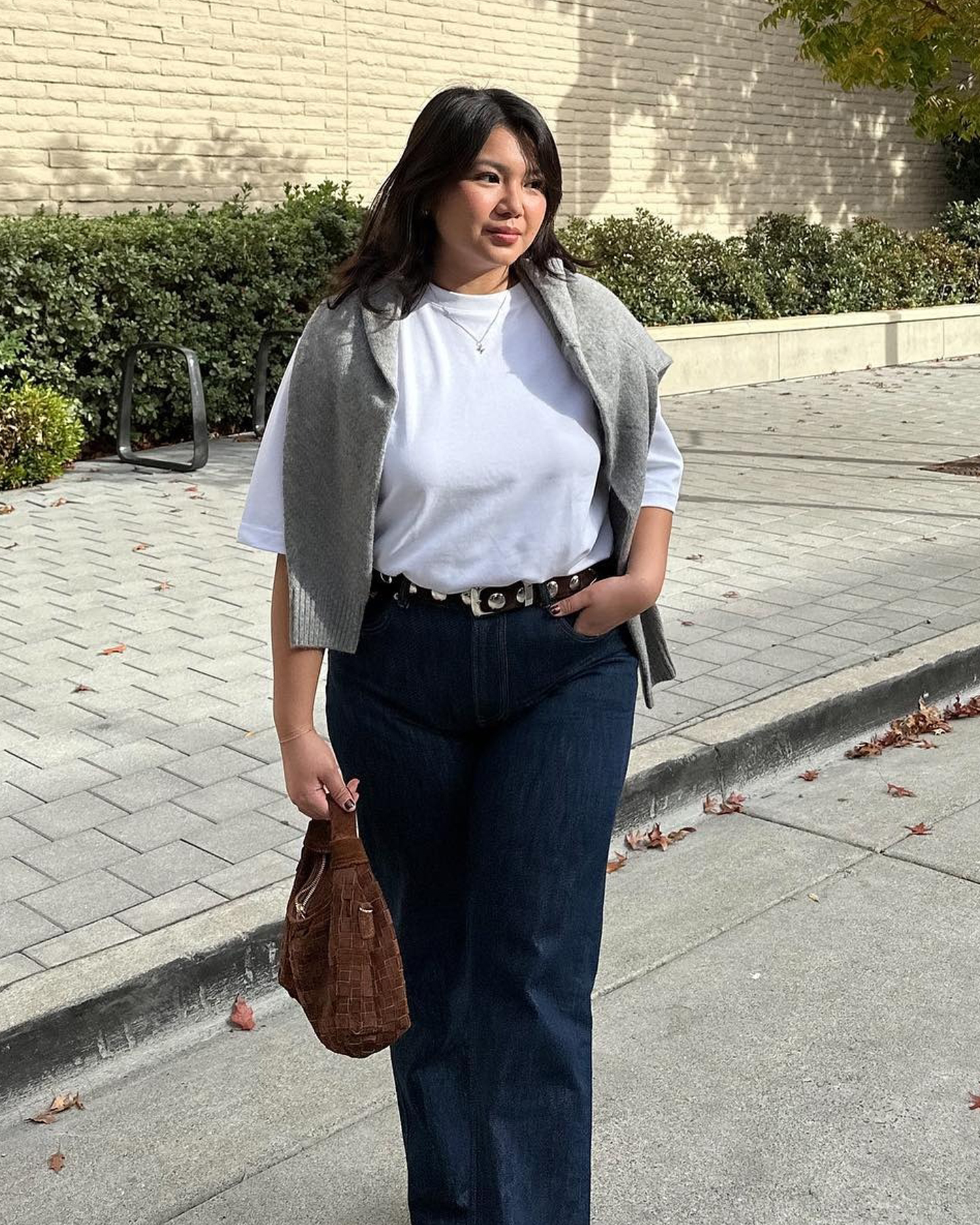 style influencer Marina Torres poses outside in a white T-shirt with a gray sweater over the shoulders, black belt with silver studs, a brown woven suede bag, and dark-wash jeans