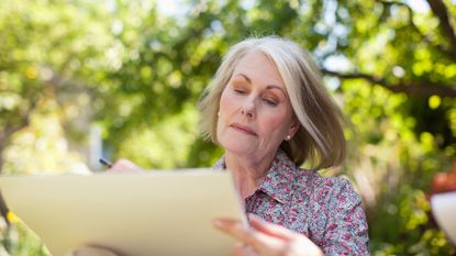 An older woman focuses on sketching while sitting outside.