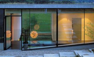 Outside image looking through the glass wall of windows and door frame, into The Biscuit House by AU*M Architectes Urbanistes, Lyon, stone steps, gravel pathway, potted plants, yellow interior lighting