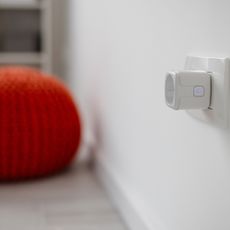 Smart plug in wall socket, with a burnt orange knitted pouffe and a plant in the background