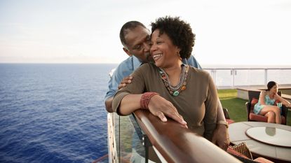A middle-aged couple laugh together while standing at the rail of a large boat. 