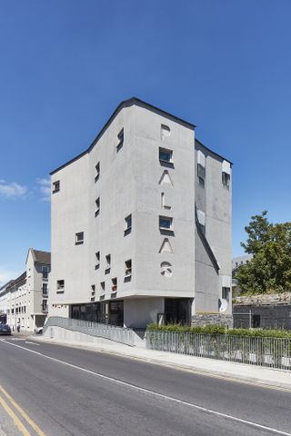 Closer exterior view of the grey concrete Pálás Cinema building under a clear blue sky. The building features multiple windows and the wording 'PÁLÁS' on the side. There are residential buildings, greenery and a road nearby