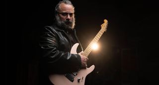 Rick Graham is photographed in a dark setting with a warm stagelight in the background; he holds his new signature electric guitar, a shell-pink S-style with a reverse headstock