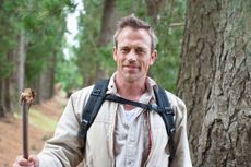 A middle aged man walks in the woods. He has a walking stick and backpack, and is smiling at the camera.