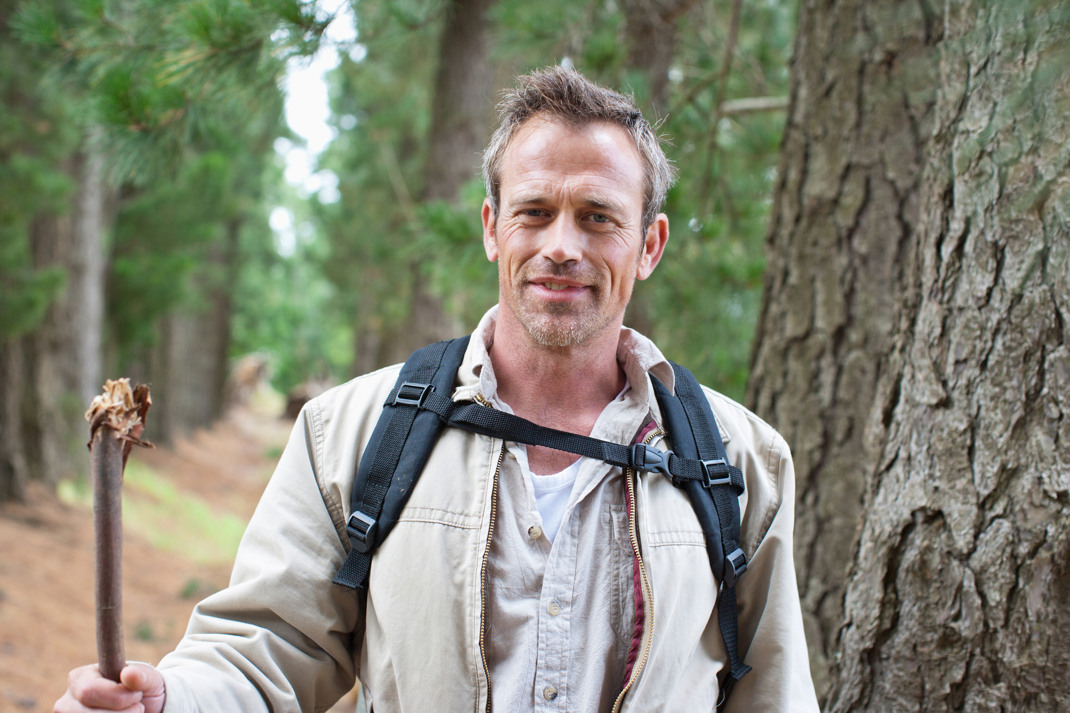 A middle aged man walks in the woods. He has a walking stick and backpack, and is smiling at the camera.