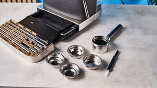 a black espresso machine by kitchenaid photographed against a blue background with silver chrome buttons and a pressure dial and steam wand and the accessories like extra single-wall and dual-wall portafilters and espresso cleaning brush