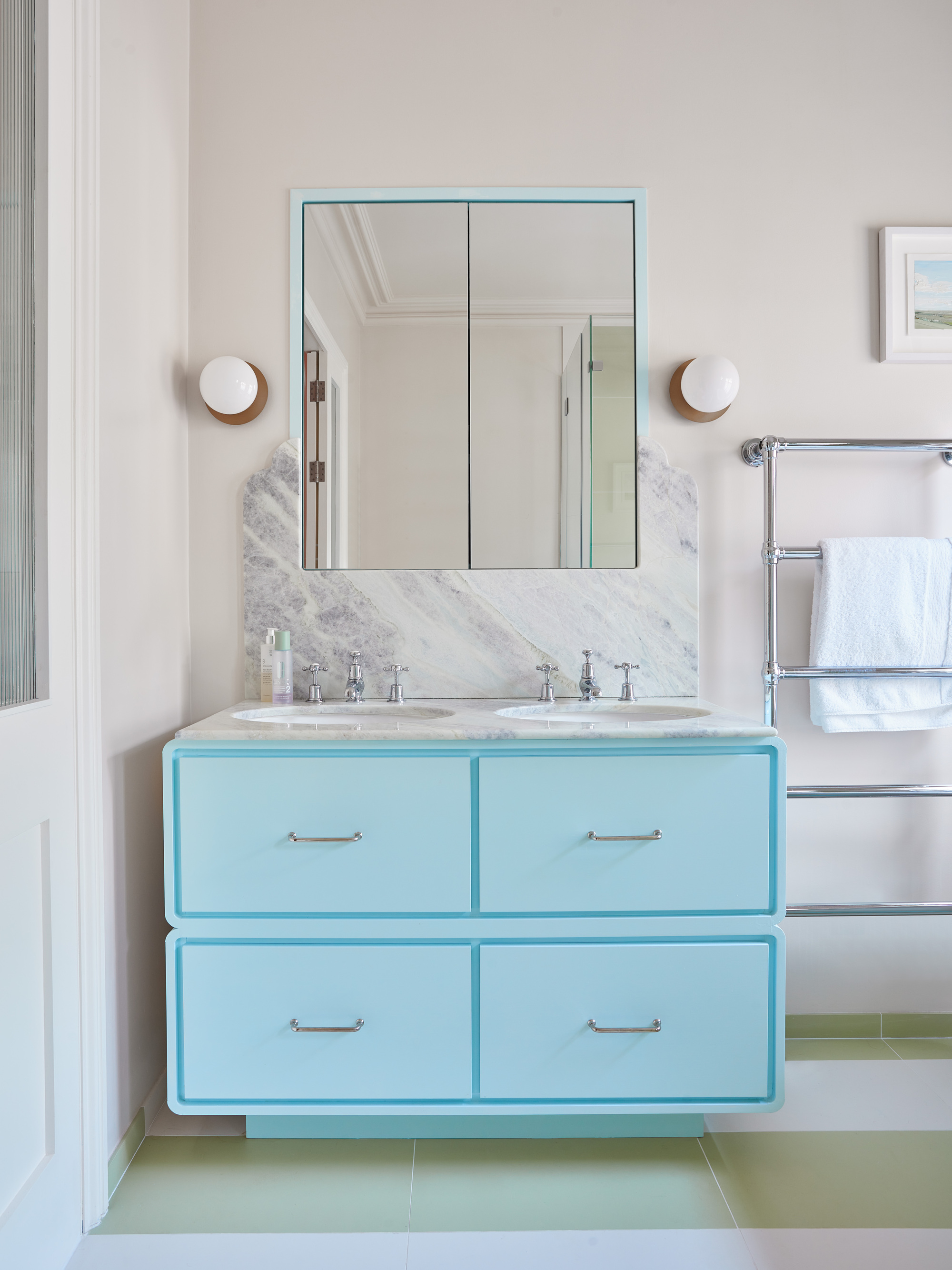 bathroom with white walls, marble backsplash, light blue floating cabinetry, two wall sconces, heated towel rack, green and white striped floor tiles