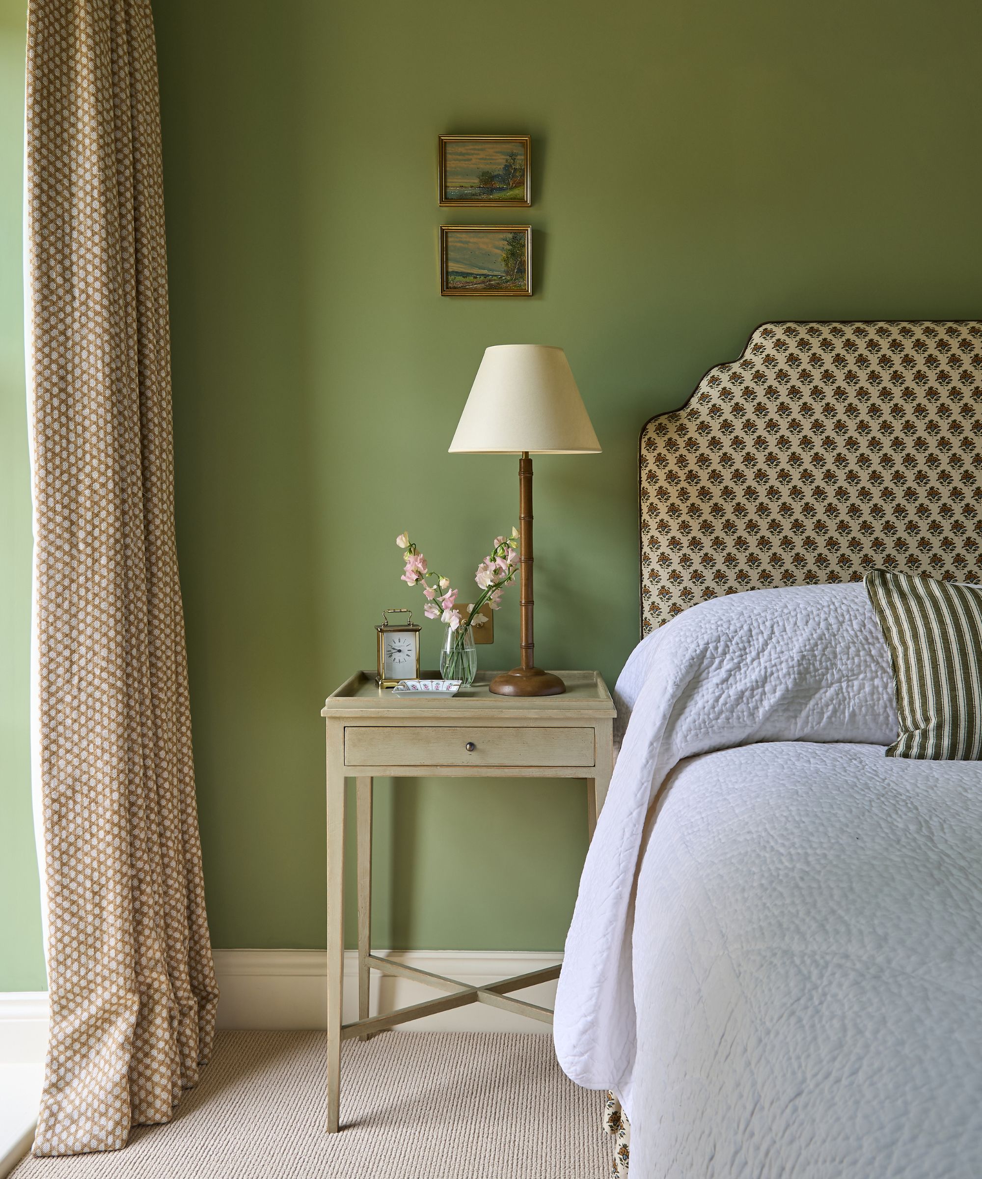 Traditional bedroom with soft olive-green walls, a patterned upholstered headboard, and a pale wood bedside table styled with fresh flowers and a classic table lamp.