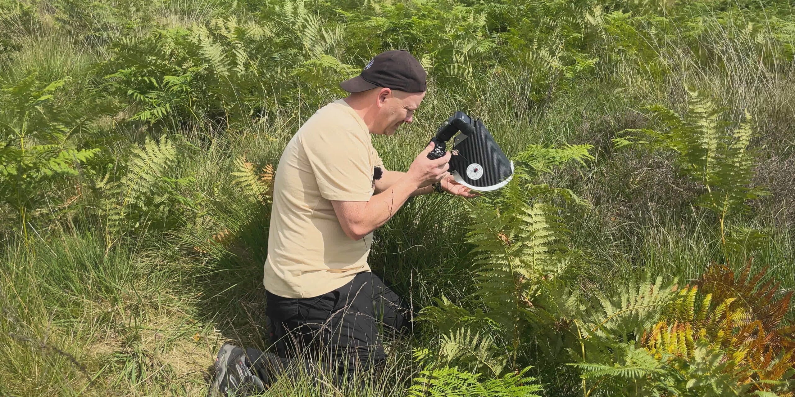 A person kneels among ferns, holding a device aimed at the ground, appearing to engage in close observation or investigation