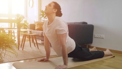 man wearing a white tshirt performing an extended spinal stretch from an exercise mat on the floor. he's sideways to the camera and there's living room furniture behind him with sunlight shining through.