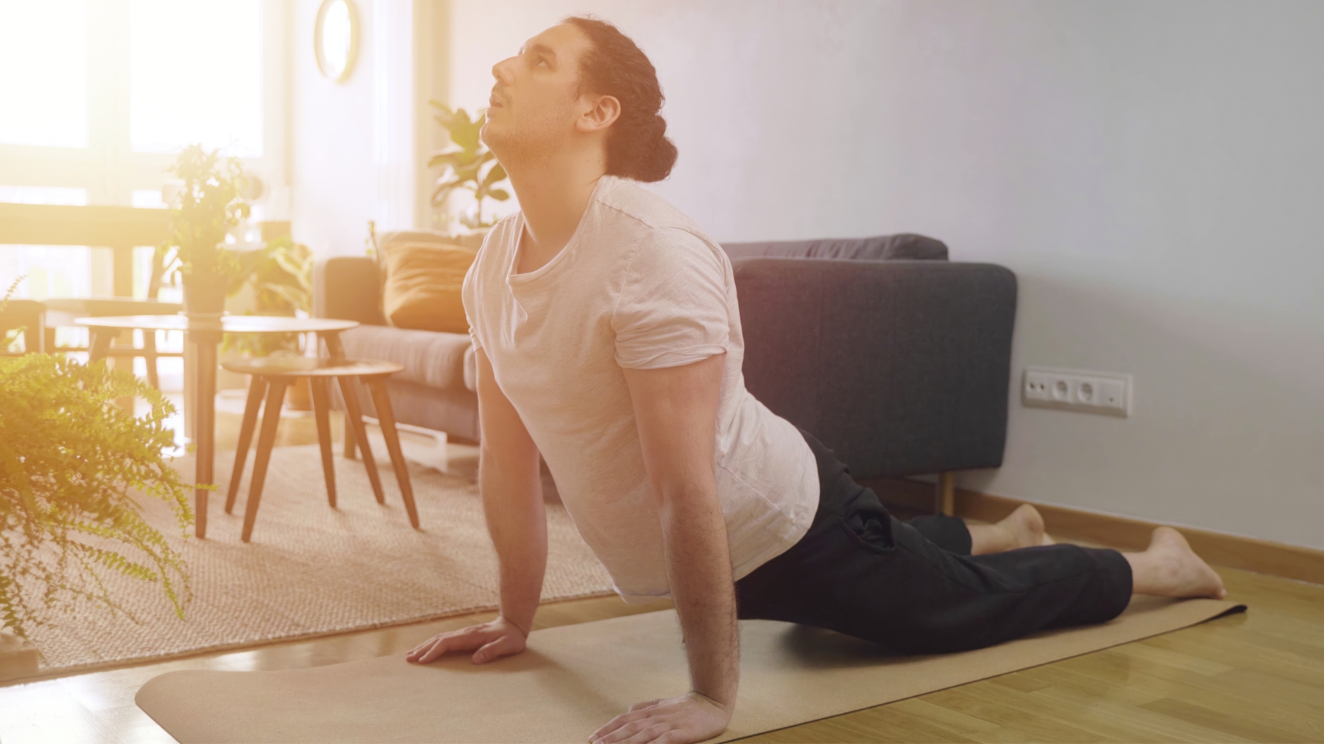 man wearing a white tshirt performing an extended spinal stretch from an exercise mat on the floor. he&#039;s sideways to the camera and there&#039;s living room furniture behind him with sunlight shining through.