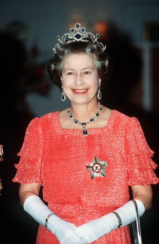 Queen Elizabeth wears a shimmery red dress with white gloves, a tiara, and the George VII Sapphires