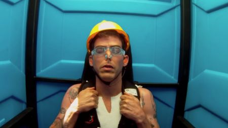 Steve-O wearing a yellow hard hat with goggles on and a nose plug, with his eyes closed, prepared for the portapotty stunt. 
