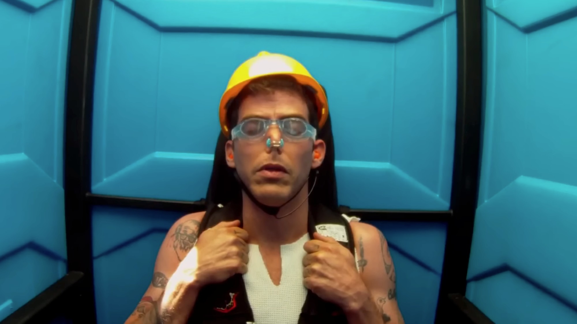 Steve-O wearing a yellow hard hat with goggles on and a nose plug, with his eyes closed, prepared for the portapotty stunt. 