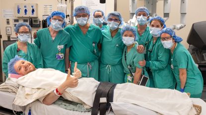 A patient is shown in the foreground of the image lying down on a bed in the operating theatre. He is wearing a blue surgical hair net and is covered by a white sheet. He is smiling at the camera with his thumbs up. Behind the bed is a group of medical professionals all dressed in blue scrubs, with blue surgical face masks and hair nets.