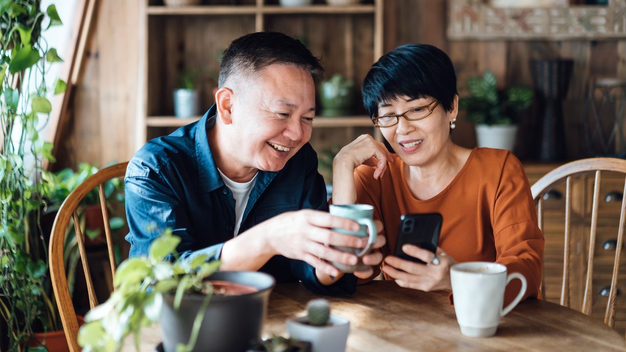 A couple smile as they sit together at a table and look at her smartphone. 